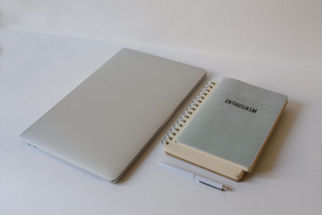 A minimalist still life photo of a closed laptop, a notepad labeled 'Enthusiasm', and a pen.